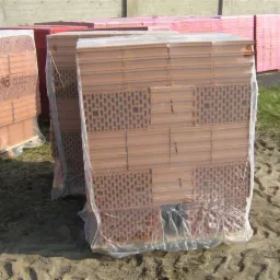 Palety z czerwonymi bloczkami ceramicznymi Porotherm, owinięte folią, składowane na zewnątrz na placu budowy, widoczne ślady opon na ziemi.